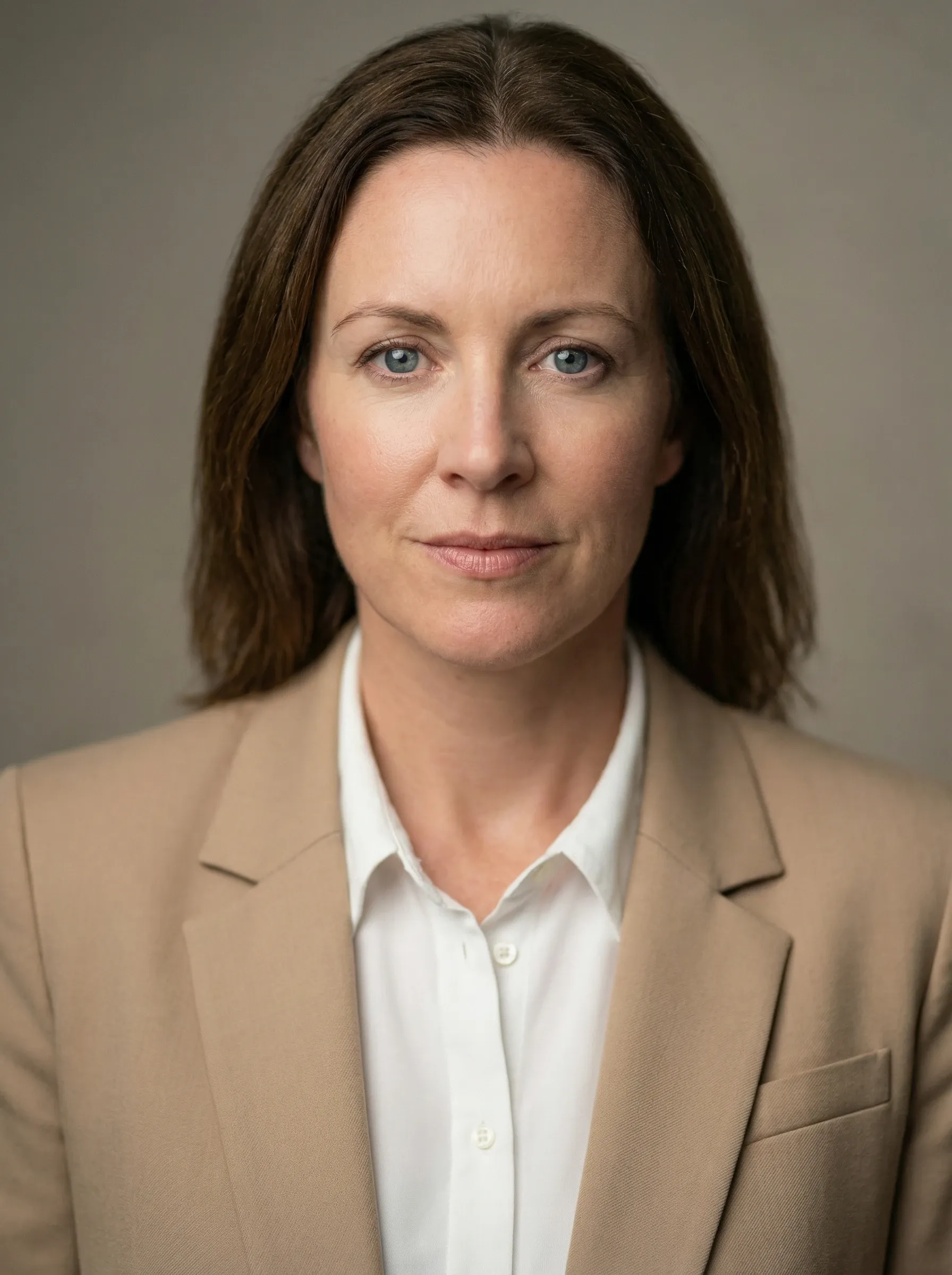 Professional headshot of a woman in a beige blazer and white shirt against a gray studio background